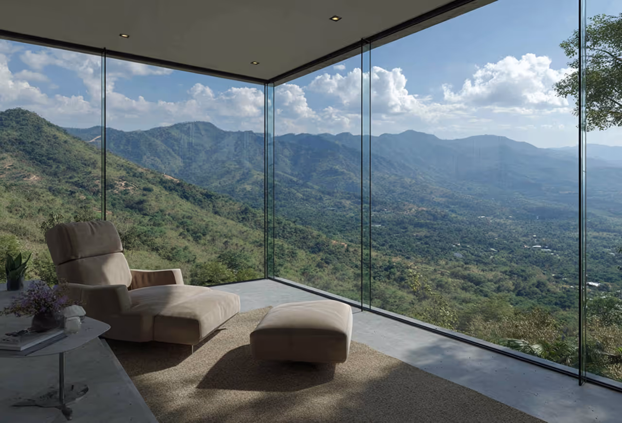 Modern lounge chair with ottoman inside a room with floor-to-ceiling glass windows overlooking green mountain landscape under a partly cloudy blue sky.